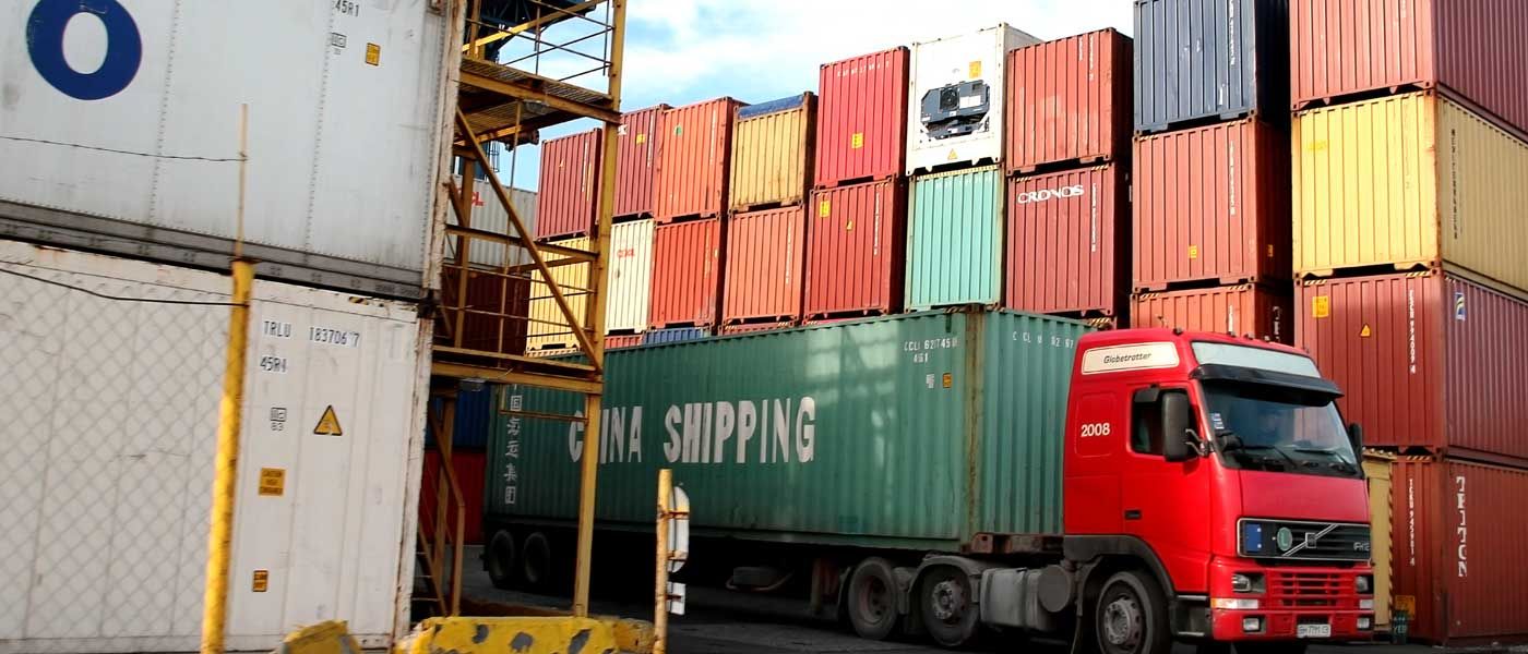 A truck carrying a China Shipping container in front of a stack of shipping containers